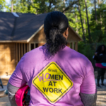Een Habitatvrijwilliger staat op de bouwplaats met een roze veiligheidshelm onder haar arm. Ze draagt een paars t-shirt met de tekst: (wo)men at work.